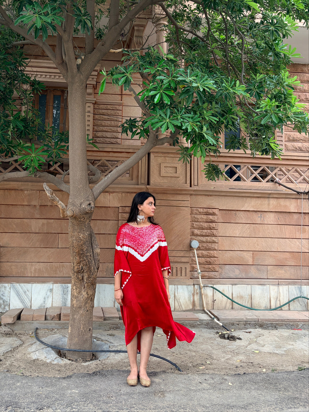 A woman wearing a vibrant red dress with white shibori hand tie-dye and a matching red headpiece stands under a large tree in front of a stone building.