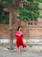 A woman wearing a vibrant red dress with white shibori hand tie-dye and a matching red headpiece stands under a large tree in front of a stone building.